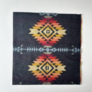 May include: A square fabric swatch featuring a dark charcoal background with two symmetrical geometric diamond motifs. The patterns incorporate gradients of red, orange, yellow, and cream, separated by a horizontal light blue arrow-like line.