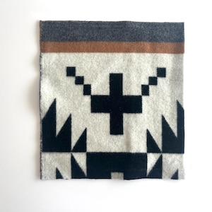 May include: A square, patterned textile featuring a geometric design. The top edge has a dark gray band above a brown stripe. The main field is off-white with black geometric shapes, including a cross and triangles. The textile has a soft, textured appearance.