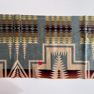 May include: Rectangular blanket with a geometric pattern in shades of blue, brown, beige, yellow, and burgundy. The blanket has a brown border and a red accent. The design features triangles, diamonds, and stepped patterns.