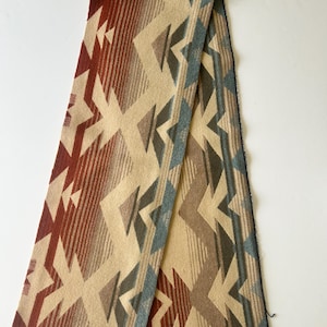 May include: A long rectangular scarf featuring a geometric pattern in shades of cream, rust red, brown, and muted blue. The fabric displays a repeating angular design with gradient color transitions and a soft, woven texture throughout the piece.