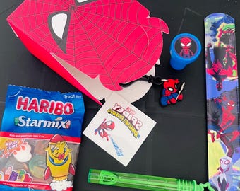 May include: A collection of Spider-Man themed party favors. Includes a red and white Spider-Man mask-shaped box, a bag of Haribo StarMix candy, a blue stamp, a keychain, a temporary tattoo, and a green bubble wand. The items are arranged on a black surface.