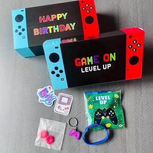 May include: Two black boxes designed as game consoles, one with "HAPPY BIRTHAY" and the other with "GAME ON LEVEL UP" in colourful letters. Accessories include stickers, a keyring, a bracelet, and small bags with pink items.