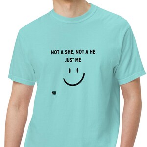 Puede incluir: Una camiseta azul claro con el texto "NOT A SHE, NOT A HE JUST ME" sobre un gráfico de una cara sonriente. Las iniciales "NB" están impresas debajo del gráfico. La camiseta está hecha de un material suave.