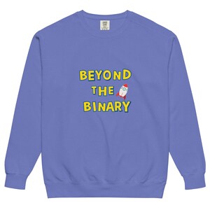 Puede incluir: Sudadera azul bígaro con el texto "BEYOND THE BINARY" en letras amarillas con contorno azul. Un gráfico de cohete de dibujos animados está al lado del texto. La sudadera tiene mangas largas y cuello redondo.