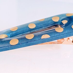 May include: A teal blue hair clip with gold polka dots and a rose gold metal clasp.