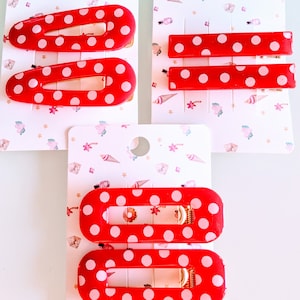 May include: Red hair clips with white polka dots are displayed on decorative cards. The clips come in various shapes, including oval and rectangular, and feature gold-colored metal clasps. The cards have a pastel pattern.