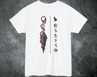 Puede incluir: Una camiseta blanca de manga corta con un gráfico de una daga kunai gris envuelta en un cordón rojo. Junto a la daga hay una silueta de un pequeño pájaro negro y el texto japonés 影を生きる命, que significa vida viviendo en las sombras.