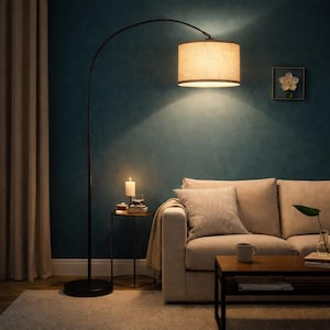 May include: A modern arc floor lamp with a beige drum shade illuminates a living room. The black metal lamp stands tall, casting a warm glow over a beige sofa, coffee table, and side table with a lit candle.