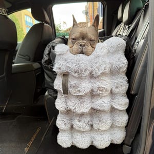 May include: A French Bulldog sits in a car seat carrier. The dog is in a fluffy, light grey pet carrier with a layered, cloud-like design. The carrier is secured with a black strap. The car's interior is visible in the background.