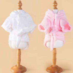 May include: Two plush hooded robes displayed on wooden mannequins. One robe is solid white and the other is solid light pink. Both robes feature short sleeves, a front button closure, and a matching fabric tie belt cinched at the waist.