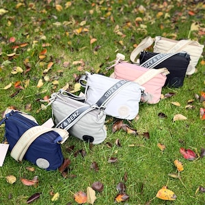 May include: A collection of crossbody bags in various colors, including navy blue, light gray, pink, black, and beige, are arranged on a bed of green grass and fallen leaves. Each bag features a matching strap with the brand name "SUZY & BENZ" printed on it.