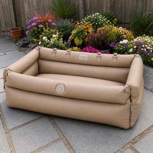 May include: A rectangular beige pet bed with high, padded walls and a smooth, leather-like finish. The bed features a central circular logo on the front panel, a small rectangular brand label on the back interior wall, and metal zippers on the sides.