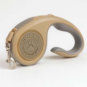 Könnte beinhalten: Einziehleine für Hunde in Beige und Grau. Die Leine hat ein beigefarbenes Gehäuse mit einem grauen Griff und Akzenten. In der Mitte der Leine steht "SUZY & BENZ LONDON BOUTIQUE" um einen Pfotenabdruck herum.