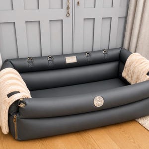 May include: A rectangular dark gray pet bed with high padded sides, featuring a beige faux fur throw draped over each end. The bed includes gold-tone metal D-rings along the inner rim, side zippers, and a circular logo patch reading Buster and Punch London.