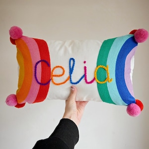 May include: A rectangular white throw pillow featuring a rainbow pattern on the left and right sides. The name Celia is embroidered in colorful yarn across the center. The pillow is accented with pink pom-poms at each of the four corners.