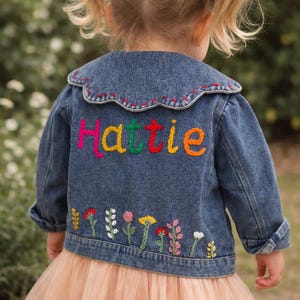 May include: A blue denim jacket featuring a scalloped collar with red stitching, the name Hattie embroidered in multicolored letters across the back, and a row of various colorful embroidered flowers along the bottom hem of the garment.