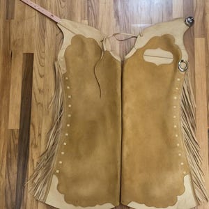 Peut inclure: Chaps en cuir de couleur beige avec une texture semblable à du daim, des bords festonnés et des franges sur les côtés. Les chaps ont des coutures décoratives et une sangle de type ceinture en haut. Un anneau et une boucle en métal sont visibles.