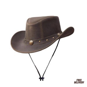 Puede incluir: Sombrero de vaquero de cuero marrón con ala ancha y copa alta. El sombrero presenta tachuelas metálicas decorativas y un medallón en la banda. Lleva una correa de barbilla negra. Estilo western clásico.