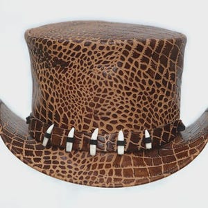 Puede incluir: Un sombrero de copa marrón con un patrón texturizado similar al de un cocodrilo. El sombrero presenta un ala curvada y una banda adornada con detalles blancos y negros. El diseño del sombrero recuerda a un estilo vintage.