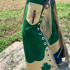Puede incluir: Chaps de cuero de gamuza verde y beige con flecos y botones plateados. Los chaps presentan un diseño de trébol y un bolsillo con una correa de cuero. Un artículo de estilo western.