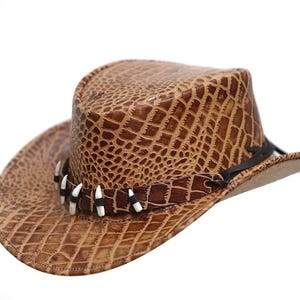 Puede incluir: Sombrero de vaquero marrón con un patrón texturizado similar al de un cocodrilo. El sombrero presenta una banda decorativa con detalles blancos y negros y una correa de barbilla negra. Accesorio de estilo western.