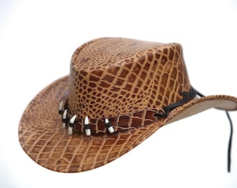 May include: Brown cowboy hat with a textured, crocodile-like pattern. The hat features a decorative band with white and black accents and a black chin strap. The hat is a western-style accessory.