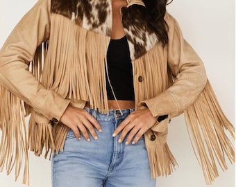 Könnte beinhalten: Beigefarbene Wildlederjacke mit langen Fransen und einem Kuhfell-Joch. Die Jacke hat eine Knopfleiste und lange Ärmel. Das Model trägt eine blaue Jeans und ein schwarzes Top. Western-Stil.