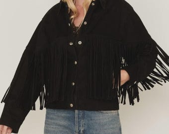 May include: Black denim jacket with long fringe detailing across the chest and sleeves. The jacket features a button-down front and a classic collar. The fringe adds a bohemian flair to the jacket's design. The jacket is paired with blue jeans.