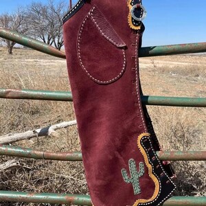 Peut inclure: Chaps en daim bordeaux avec garniture noire et jaune, une application de cactus et une poche en cuir. Les chaps sont suspendus à une clôture métallique dans un cadre rural. Boucle argentée et lanière en cuir.