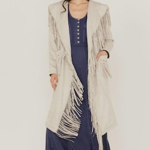 Può includere: Un lungo cappotto beige effetto scamosciato con lunghe frange sulle maniche, sul davanti e sull'orlo. Il cappotto è aperto, rivelando un vestito blu navy sottostante. Il cappotto ha un design classico di ispirazione western.
