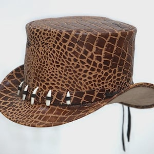 Puede incluir: Un sombrero de copa de cuero marrón con un patrón de piel de cocodrilo. El sombrero presenta un ala ancha y está adornado con detalles blancos y negros. Cuerdas negras cuelgan del sombrero.