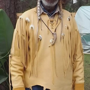 Puede incluir: Una camisa de ante de color beige claro con flecos y botones decorativos. La camisa tiene un dobladillo rasgado y se usa con vaqueros azules. También se ven un sombrero negro y un collar con un colgante.