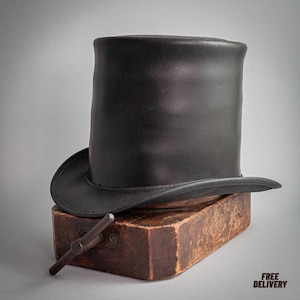 May include: A black leather top hat with a rounded crown and a curved brim. The hat rests on a wooden hat block with a metal tool. The image has a neutral background and the words "FREE DELIVERY" are in the bottom right corner.