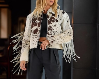 May include: A cropped jacket with a cowhide pattern in brown and white. The jacket features a collar, a zipper closure, and fringe detailing on the sleeves. The model is wearing black pants and a black top.