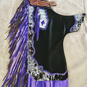 Könnte beinhalten: Schwarze Lederchaps mit lila Fransen und Akzenten. Die Chaps haben ein Kuhfellmuster, Spielkartendetails und braune Lederakzente. Das Design umfasst eine dekorative Oberseite mit einem Riemen.