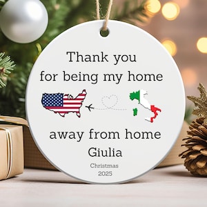 May include: A white ceramic Christmas ornament with a rope hanger. The ornament features the text "Thank you for being my home away from home Giulia Christmas 2025." It includes a map of the USA and Italy, connected by a dotted line and airplane.