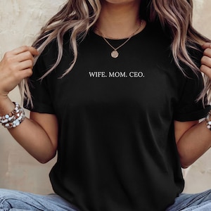 May include: A black short-sleeved t-shirt featuring the white text WIFE. MOM. CEO. printed across the chest. The shirt has a crew neckline and is worn with blue denim jeans, layered gold necklaces, and beaded bracelets on both wrists.