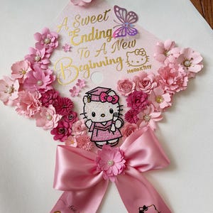 May include: A pink graduation cap adorned with pink paper flowers, a butterfly, and a large pink bow. The cap features the text "A Sweet Ending To A New Beginning" and a Hello Kitty graphic. The ribbon says "2026".
