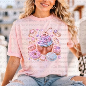 May include: A light pink t-shirt featuring a central graphic of a cupcake topped with a cherry, surrounded by various rings, macarons, and small cakes in shades of purple, pink, and blue with sparkling glitter and gemstone accents.