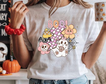 May include: White t-shirt with a colorful Halloween-themed graphic. The design includes the word "BOO!" in orange and pink, a spider, a ghost, flowers, and a candy corn hat. The shirt is paired with blue jeans.