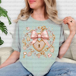May include: A light sage green t-shirt featuring a central graphic of a large, sparkling heart-shaped padlock adorned with a prominent pink bow. The design is surrounded by a circular arrangement of gold and silver chains, smaller heart-shaped padlocks, and scattered gems.