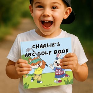 May include: A child holds a children's book titled "Charlie's ABC Golf Book." The book cover features cartoon illustrations of a fox playing golf and a rabbit. The background is a light blue with the title in black letters.