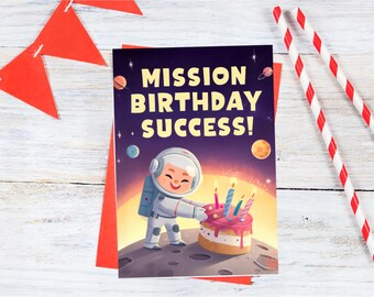 May include: A birthday card with the text "MISSION BIRTHDAY SUCCESS!" features an astronaut character and a cake with candles. The card is on a red envelope, with red and white striped straws and a red banner in the background.