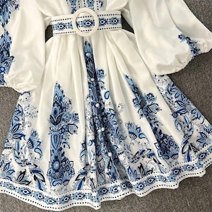 K&ouml;nnte beinhalten: Wei&szlig;es Kleid mit blauem Blumenmuster. Das Kleid hat lange, puff&auml;rmel, eine Knopfleiste vorne und einen passenden G&uuml;rtel mit einer runden Schnalle. Das blaue Design wiederholt sich an den &Auml;rmeln und am unteren Saum.