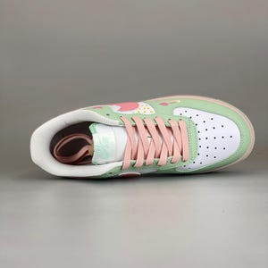 May include: A white and green sneaker with pink laces. The shoe features a strawberry design on the upper, a white toe box with black dots, and a pink sole. The tongue has a mint green "Air" logo.
