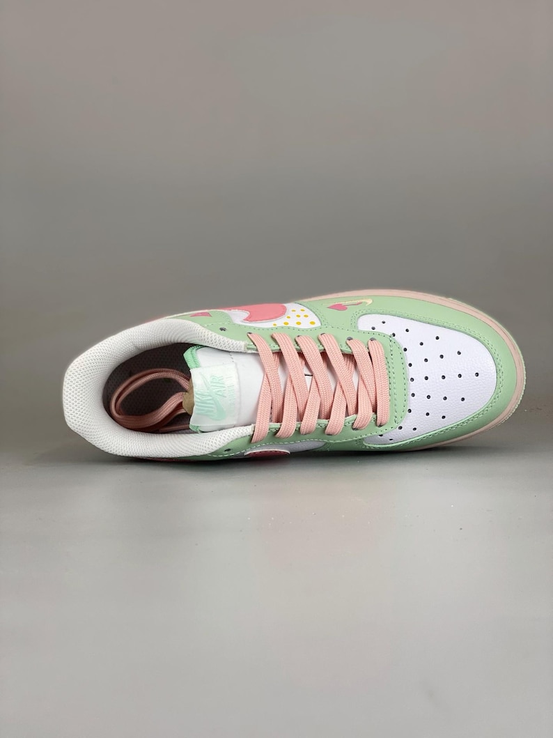 May include: A white and green sneaker with pink laces. The shoe features a strawberry design on the upper, a white toe box with black dots, and a pink sole. The tongue has a mint green "Air" logo.
