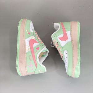 May include: Pair of mint green, white, and pink Air Force 1 sneakers. The shoes feature a white base with mint green overlays and pink accents on the swoosh, laces, and sole. The design includes heart and card suit details.