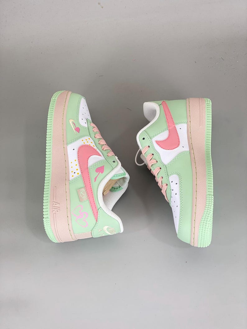 May include: Pair of mint green, white, and pink Air Force 1 sneakers. The shoes feature a white base with mint green overlays and pink accents on the swoosh, laces, and sole. The design includes heart and card suit details.