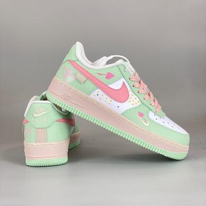 May include: A pair of pastel-colored Air Force 1 sneakers. The shoes feature a white base with mint green and pink accents. The Nike swoosh is pink, and the laces are pink. The sole is mint green and pink.