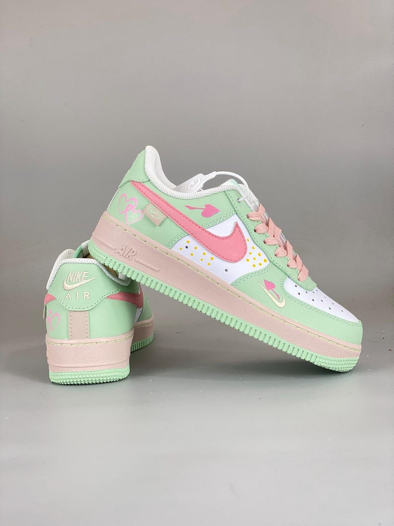 May include: A pair of pastel-colored Air Force 1 sneakers. The shoes feature a white base with mint green and pink accents. The Nike swoosh is pink, and the laces are pink. The sole is mint green and pink.